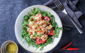 dish with green leafy salad and watermelon, topped with peeled cooked king prawns and sesame seeds. On the side there is a fork on a grey napkin, chilli peppers and olive oil dressing.